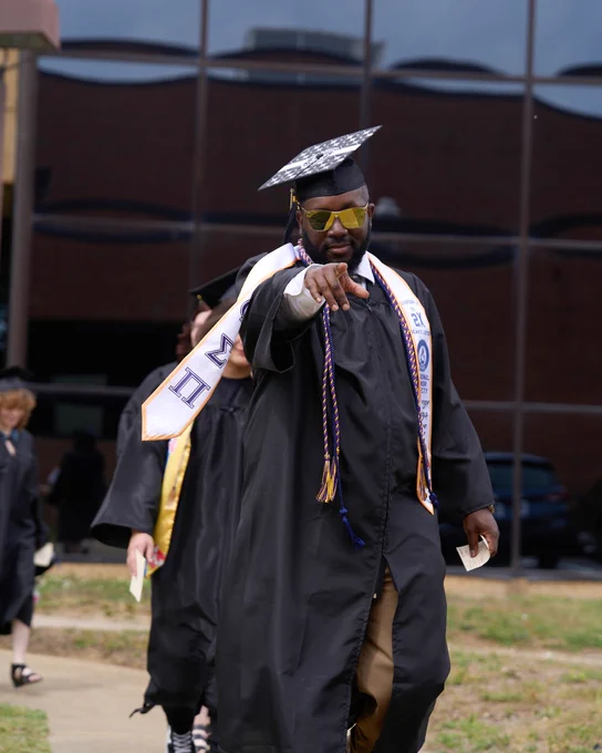 Adult male graduate wearing sunglasses, in black cap and gown pointing forward at camera walking to Chattanooga State graduation.