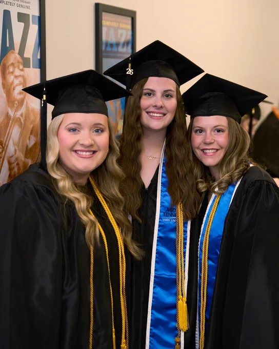 Three female graduates in black caps and gowns wearing blue stoles and gold cords posing indoors before Chattanooga State graduation.