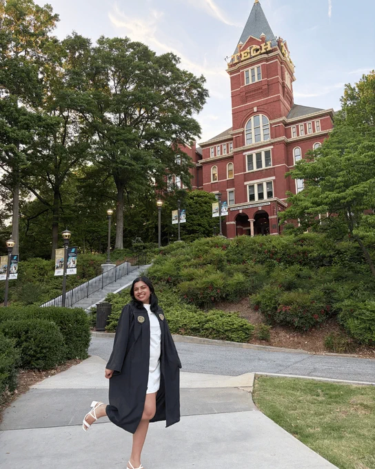 A graduate in cap and gown stands on the Georgia Tech campus with Tech Tower rising in the background, surrounded by trees and landscaped walkways.