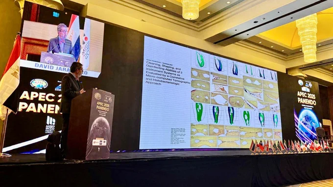 A male speaker stands at a podium on a large conference stage. A screen behind him displays a scientific slide with tooth cross‑section images and a video feed of him speaking. The stage is lined with small international flags and event banners in a conference hall.
