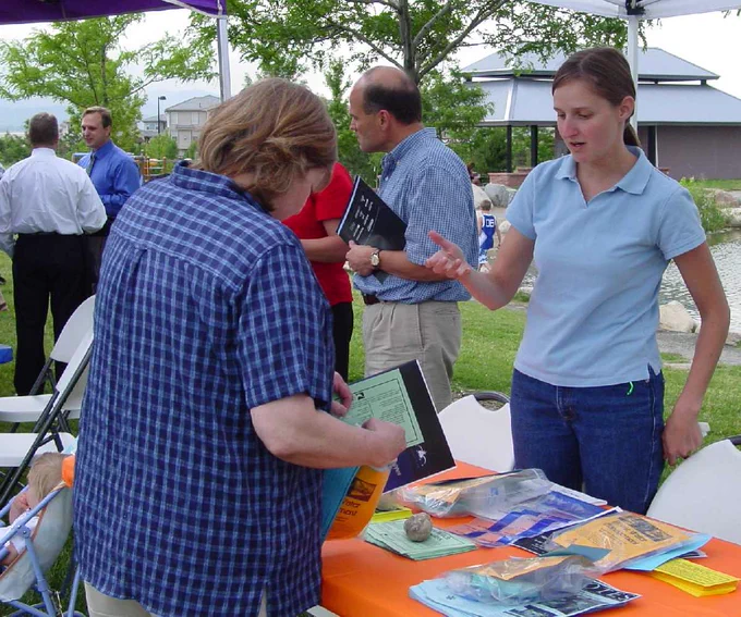 A woman at a table outdoors discusses conservation materials with another person, while others engage in conversation in the background.