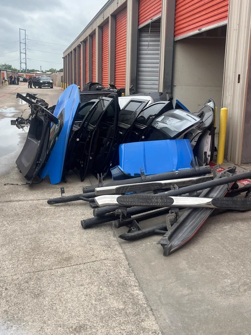 A collection of car parts, including doors and bumpers, is stacked outside metal storage units with red roll-up doors. The parts appear to be various colors, including blue and black. The scene is outdoors, and the ground is concrete.
