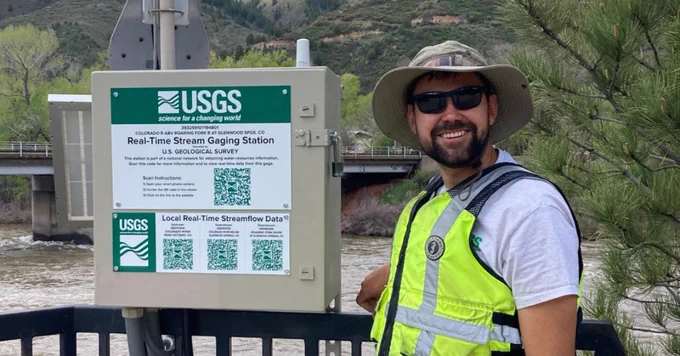 A USGS scientist standing by the Colorado River above Roaring Fork River at Glenwood Springs, CO, gage shelter.