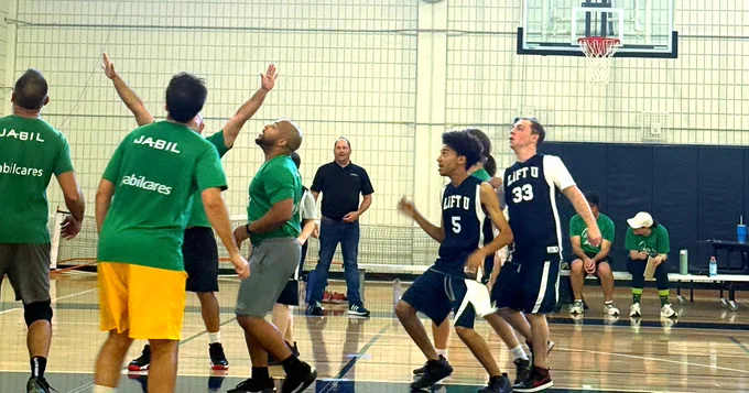 A group of individuals playing basketball in a gym. Some players are wearing green shirts with "Jabil" written on them, and others are in dark jerseys marked "Lift U." People are actively engaged in the game, with a couple of individuals sitting on the sidelines.