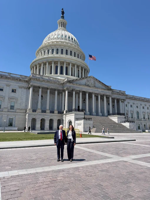 Two people stand in front of the U.S. Capitol on a sunny day, with the dome and American flag visible against a clear blue sky.