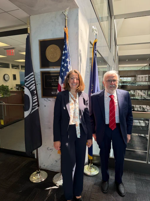 Two people stand indoors beside U.S. and POW/MIA flags and a wall plaque in a government office building.