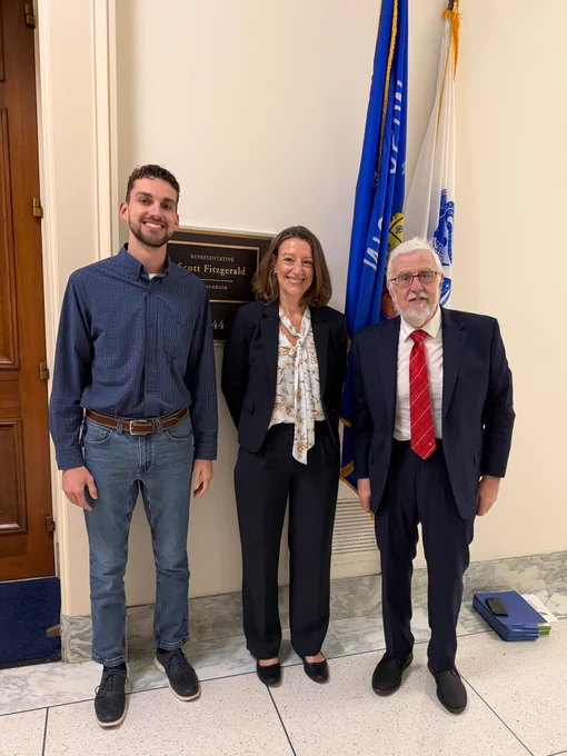 Three people stand in a hallway in front of a wall plaque and U.S. and state flags, posing for a photo inside a government building.