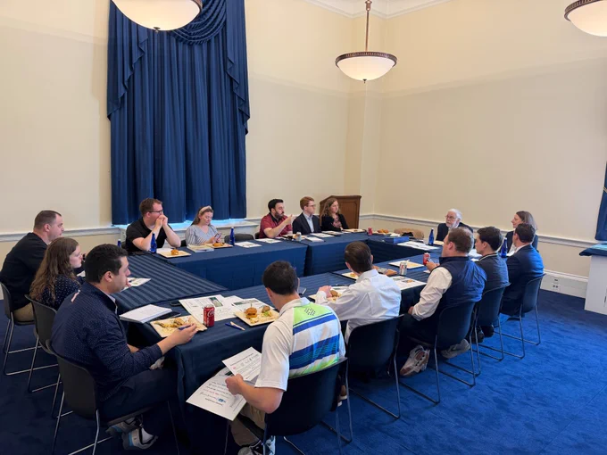 A group of people sits around a U-shaped table in a formal meeting room, eating lunch and discussing papers beneath tall curtains and hanging lights.