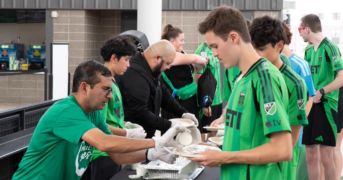A group of people, some wearing Austin FC soccer jerseys, are being served food by Jabil Cares volunteers at a counter.