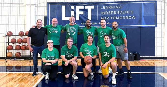 A group of people on a basketball court, some standing and some kneeling, wearing green shirts with "Jabil Cares" on them. They are holding a basketball. A "LiFT: Learning Independence for Tomorrow" banner is displayed in the background alongside basketballs on racks.