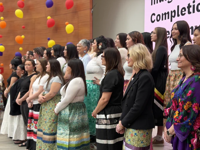 A group of people stand together, all wearing brightly colored skirts with various patterns. They appear to be part of a celebration, with colorful balloons floating in the background. A large screen displays the words "Indigenous Completion Ceremony".