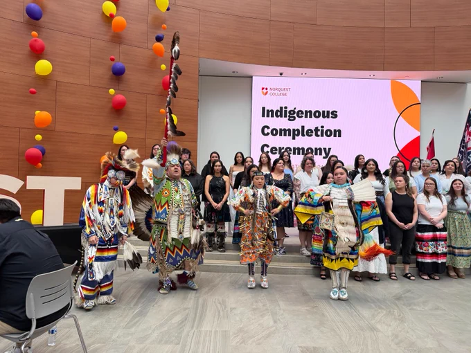 A group of Indigenous individuals in colorful traditional attire perform in front of an audience at NorQuest College's "Indigenous Completion Ceremony." Brightly colored balloons decorate the wooden-paneled room. Attendees stand near a large display screen showing the event's name.