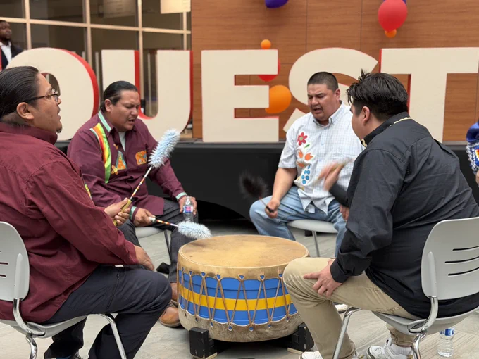 A group of people seated in a circle playing a large drum with drumsticks. The setting appears to be an indoor event or celebration, with several balloons visible in the background and large letters spelling out "NORQUEST".