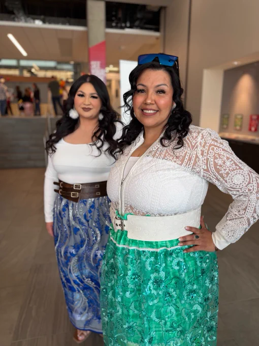 Two people smiling indoors, dressed in colorful clothing with intricate patterns. They are standing in a modern building with stairs and a lobby area visible in the background.