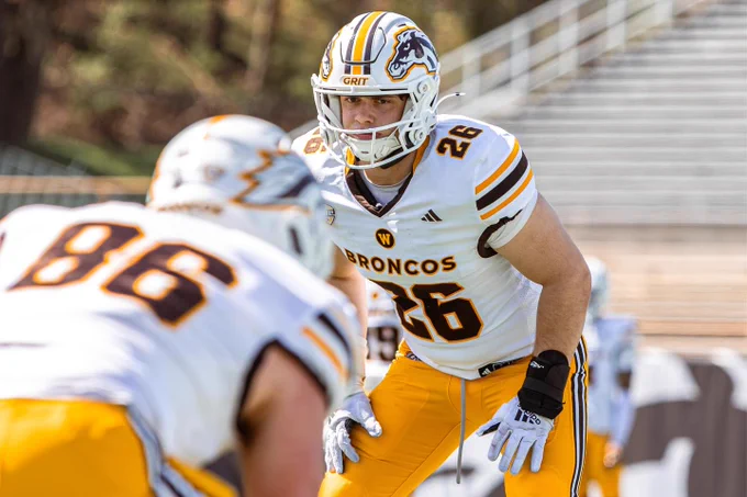 A Western Michigan football player in a white and gold uniform, numbered 26, is focused and ready on the field. Another player, with number 86, is in the foreground. 