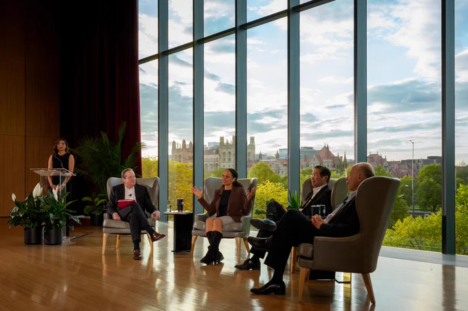A panel discussion is in progress, featuring four individuals seated in armchairs against a backdrop of a large window revealing a cityscape. A woman stands at a podium to the left. Everyone is formally dressed.