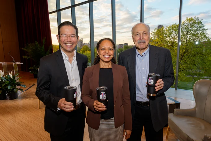 Three smiling individuals, two men and one woman, pose indoors holding \"UChicago BIG BRAINS Podcast\" cups. A large window behind them offers a view of trees and a building.
