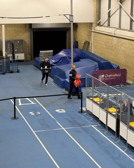 Two people entering a sports hall. One carries a ballot box, the other a red bag.