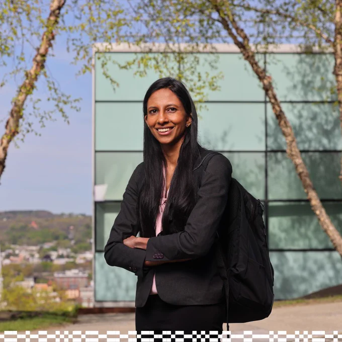 A person with long hair stands outdoors with arms crossed, wearing a blazer and holding a backpack. Trees and a modern building with reflective glass panels are in the background, with a distant view of green hills under a blue sky.