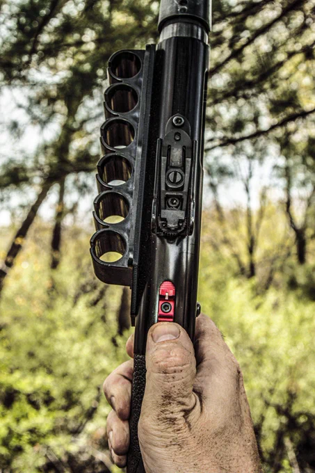Close-up outdoor view of a Mossberg shotgun equipped with the red NDZ Sight Integrated Safety Selector. A hand grips the shotgun while the upgraded slim-profile safety and side saddle shell carrier are shown in detail against a wooded background.