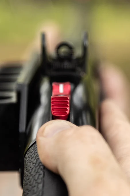 First-person close-up of the red NDZ Mossberg Sight Integrated Safety Selector aligned beneath a rear sight. The textured aluminum safety is shown in use with a thumb engaging the control for faster, more ergonomic manipulation.