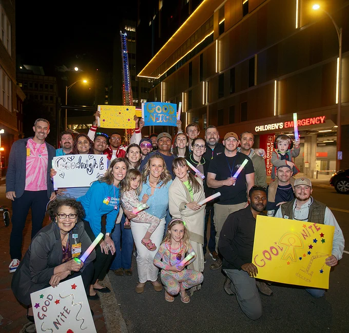 A diverse group of people stands outside at night in front of a building with a sign that reads "Children's Emergency." Some hold colorful signs and glow sticks, including one sign that says "Good Night." The group appears cheerful and united. 