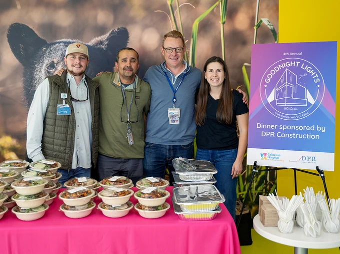 Four people pose before distributing food to team members at Children's Hospital of Richmond at VCU. A nearby sign reads: "Goodnight Lights. Dinner sponsored by DPR Construction."