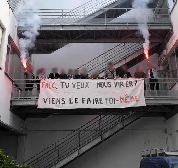 Une photo de manifestants avec des fumigènes sur une passerelle, leurs visages sont floutés. Iels tiennent une banderole avec écrit en majuscule "FALC TU VEUX NOUS VIRER ? VIENS LE FAIRE TOI-MÊME"