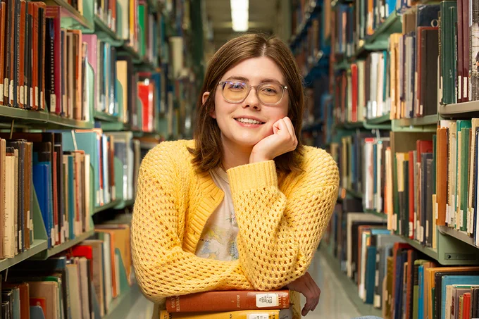 A person wearing glasses and a yellow sweater is sitting among bookshelves filled with books in a library, resting their head on their hand with a gentle smile.