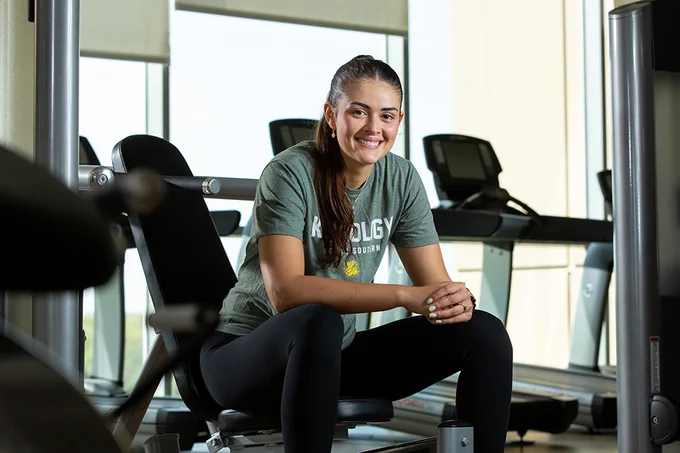 A person sitting on a gym bench, smiling, wearing a green "Kinesiology" shirt and black pants. Treadmills are visible in the background.