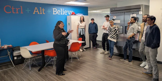 A woman addresses a group of six people standing in a modern office kitchen area with a blue wall that reads 'Ctrl   Alt   Believe'.