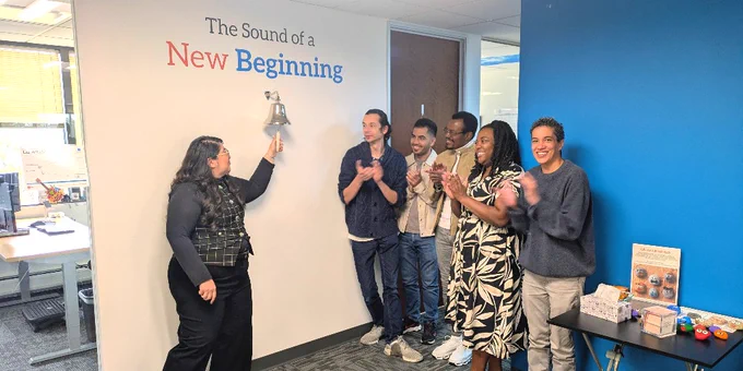 A woman rings a bell on a wall with the phrase 'The Sound of a New Beginning' while five people applaud in an office setting.
