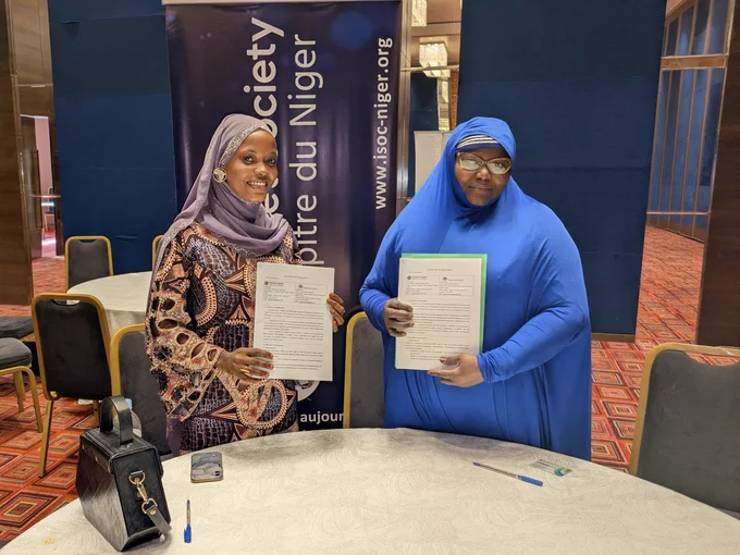 Two woman stand together and hold documents in front of an Internet Society Niger Chapter sign
