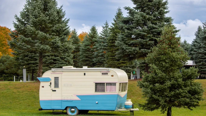 A vintage camper trailer with a white and blue exterior is parked on a grassy field. The area is surrounded by tall evergreen trees, with a backdrop of autumn-colored foliage. The sky is overcast, suggesting a cloudy day.