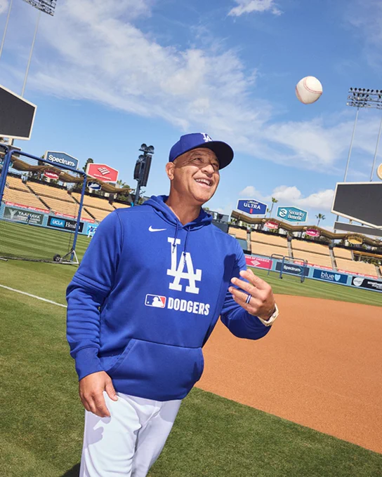 Dodgers coach Dave Roberts on a baseball field in a Los Angeles Dodgers hoodie and cap is smiling while tossing a baseball. Stadium seats and advertising boards are visible in the background under a clear sky.