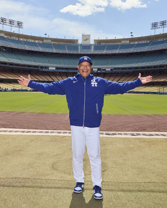 Dodgers coach Dave Roberts, in a Los Angeles Dodgers jacket and cap, is standing on the field at Dodger Stadium with arms outstretched, smiling. The stadium seats and empty field are visible in the background.