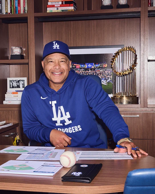 Coach Dave Roberts is seated at a desk in an office, wearing a Los Angeles Dodgers cap and hoodie. The desk has papers, a baseball, and a Dodgers notebook. Behind it is a bookshelf with various books and a photo featuring diverse flags. A championship trophy is displayed in the background.