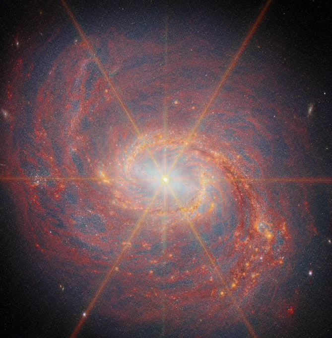 A spiral galaxy shown in infrared light. Six long and two smaller rays of light emit from the center, which are diffraction spikes created by the telescope\u2019s optics. A glowing bar spans across the center. A glittering ring of stars and dust surrounds the bar; at each side, the ring splits off into a spiral arm that winds outwards. Faint, dark red dust clouds swirl throughout the rest of the disc, backed by a pale glow from all the galaxy\u2019s stars.