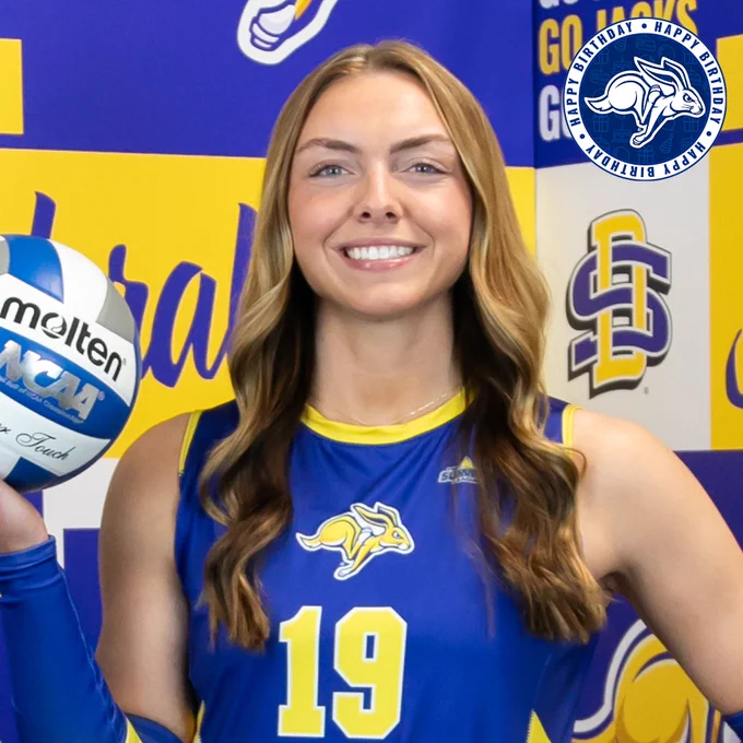 A South Dakota State volleyball athlete wearing a blue jersey with the number 19 and holding a volleyball at shoulder height. The background features a blue and yellow branded wall with Jackrabbits and SDSU logos. A circular graphic in the top corner includes the Jackrabbits logo and reads “Happy Birthday.”