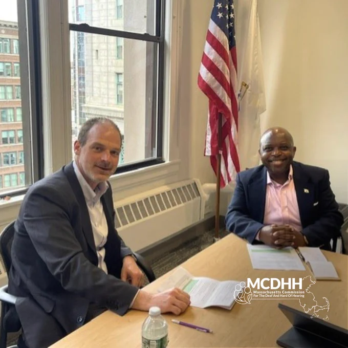 DTA Commissioner Michael Cole sits with MCDHH Commissioner Opeoluwa Sotonwa. Both men are smiling and sitting in a boardroom with an American flag in the background.