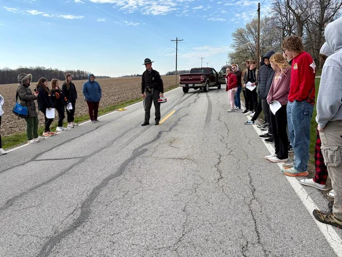 A trooper discusses crash reconstruction with students. They are standing on or alongside a road.