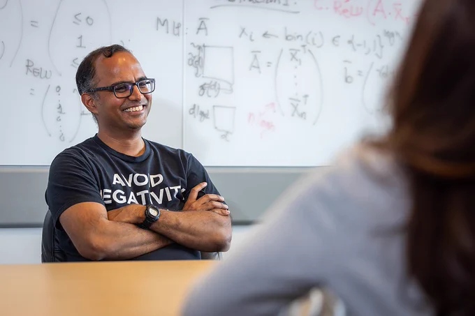 A smiling man with glasses and a \"AVOID NEGATIVITY\" t-shirt sits at a table with arms crossed. Behind him, a whiteboard is filled with equations. Another person faces away from the camera to the right.