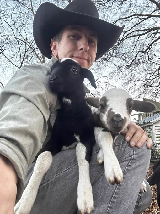 Farmer Josh Young holding two baby goats, one black and white and the other brown and white. 