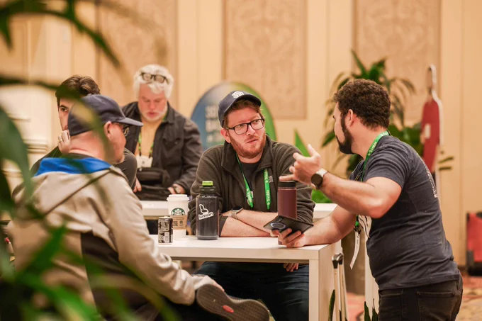 Five conference attendees chat around a high table with coffee cups and water bottles in a hotel lobby.