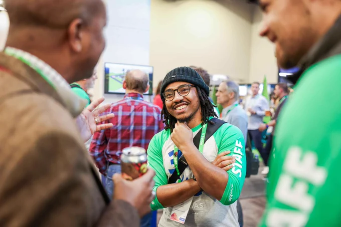 Smiling attendee in a beanie and green shirt chats with others at a busy conference or expo.