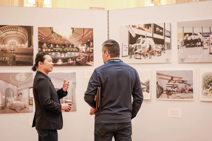 Two men talk in front of a gallery wall displaying large architectural and aerospace design images.