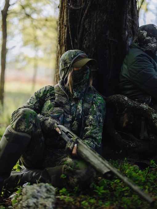 A person in camouflage gear sits against a tree, holding a shotgun, in a forest setting. Another individual in similar attire is partially visible, also leaning against the tree. Sunlight filters through the trees, creating a serene outdoor atmosphere.