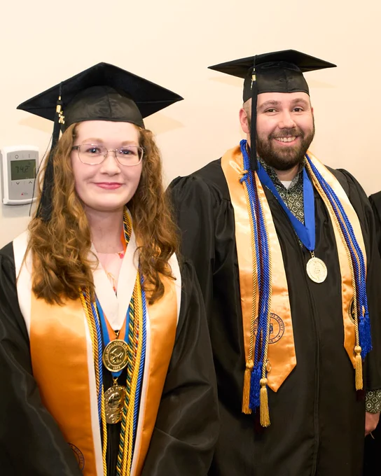 Adult male and female graduates wearing black caps and gowns with gold stoles and multiple honor cords and medals stand side by side before Chattanooga State graduation. 