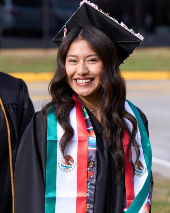 Female graduate wearing a black cap decorated with flowers and a Mexican flag stole over a black gown smiling outside before Chattanooga State graduation. 
