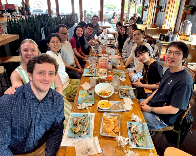 Two rows of lab members sit at a long restaurant table, which is full of eaten food and plates. They're all looking at and smiling for the camera.